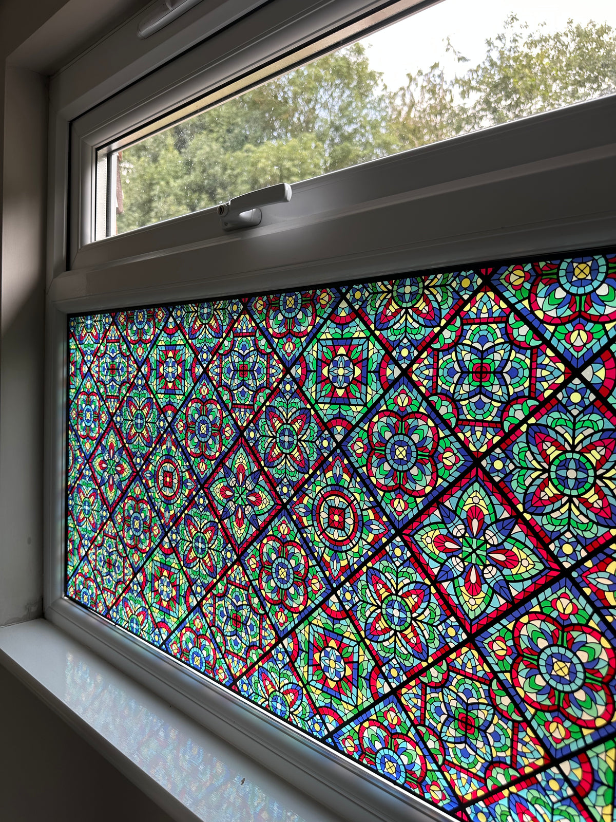 A close-up of a window with a colourful stained-glass window film covering the lower window. The pattern contains multiple bright colors including red, blue, green, yellow, and white in a repeating geometric and floral design.