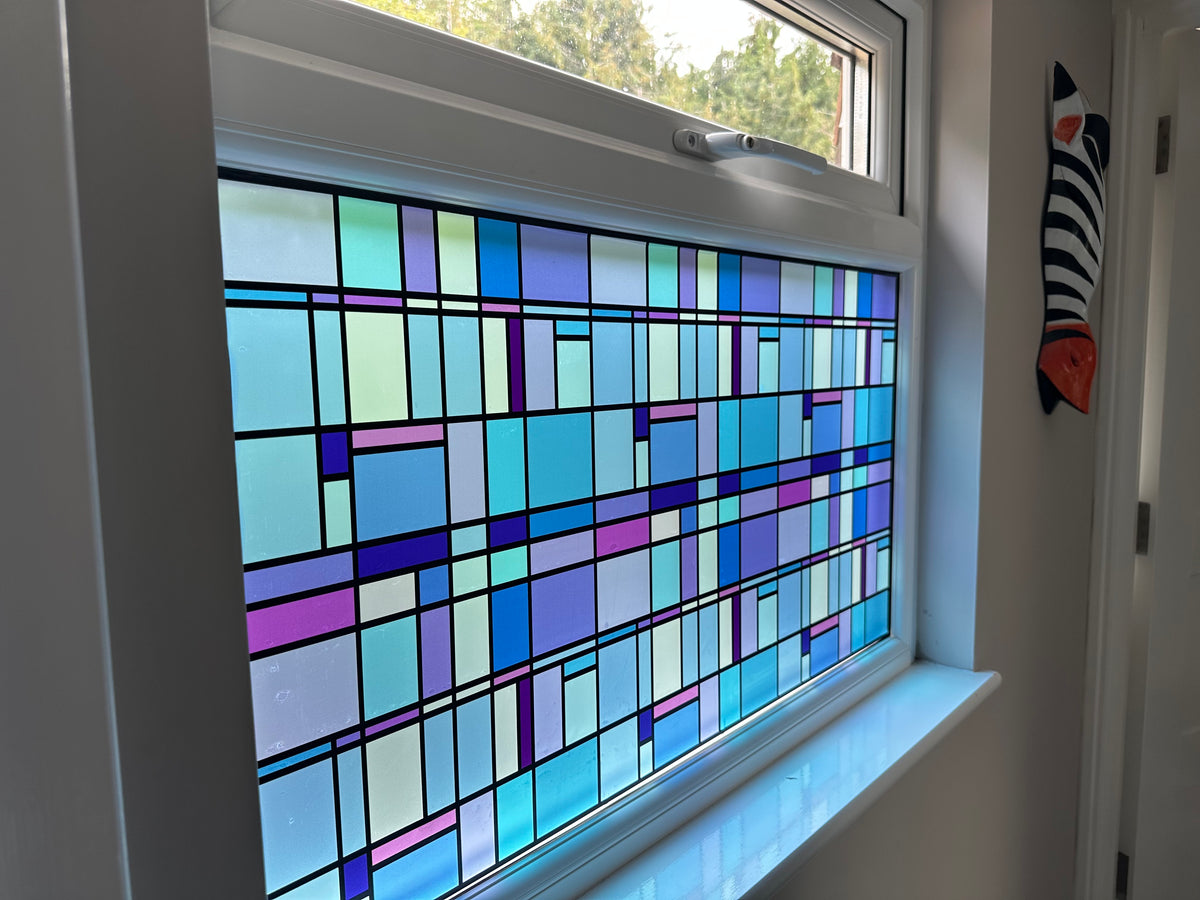 A geometric stained glass window panel with an abstract pattern of blue, purple, green, and white rectangles outlined in black, installed on a white-framed window in a home for added decor and privacy.