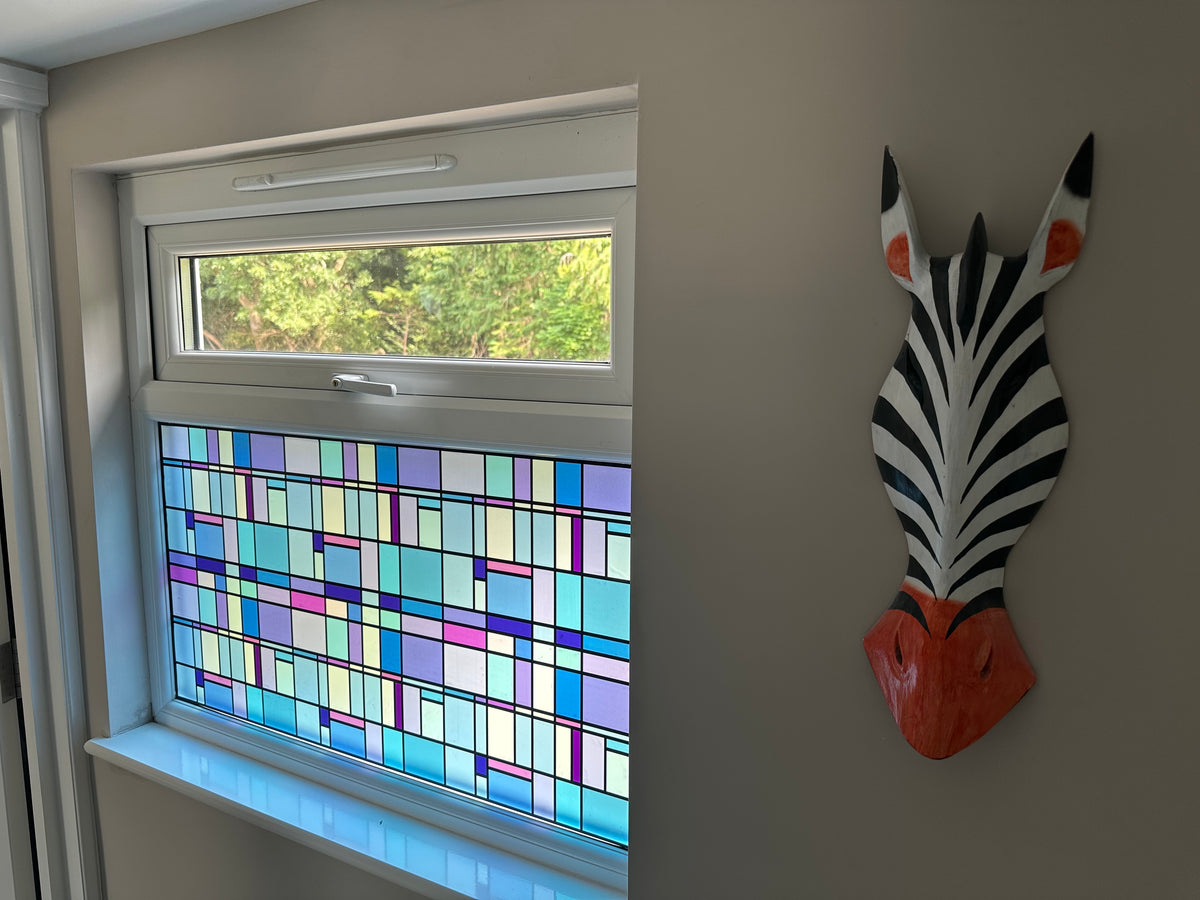A colourful stained glass window film with geometric patterns in shades of blue, purple, yellow, and green, installed in a white-framed window on a beige wall. A zebra head wall sculpture is mounted to the right.