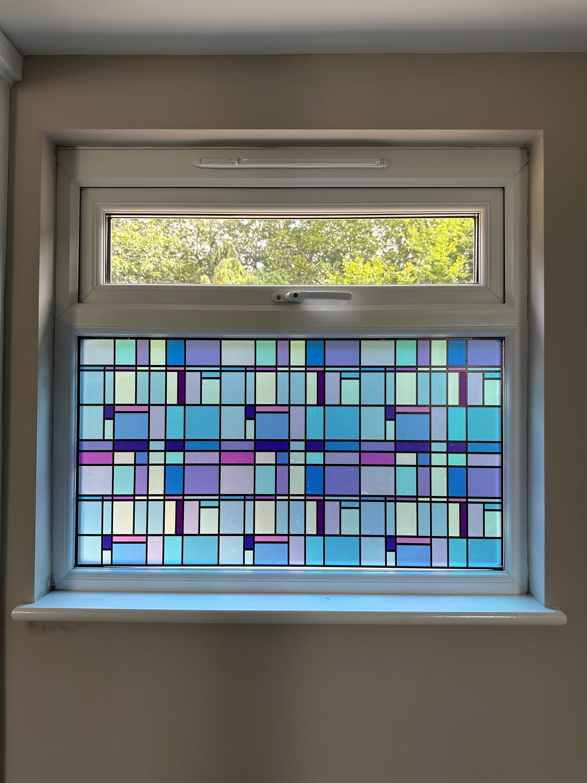 A window in a home, with colourful stained glass effect window film applied to the lower pane. The film is different shades of blue, delivering a colourful decorative upgrade and privacy solution.