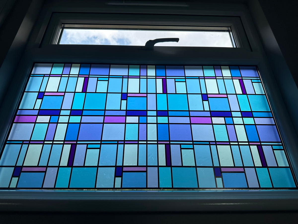 A stained glass window panel featuring a geometric pattern of blue, purple, and light blue rectangular sections divided by dark lines, set within a window in a home setting. 