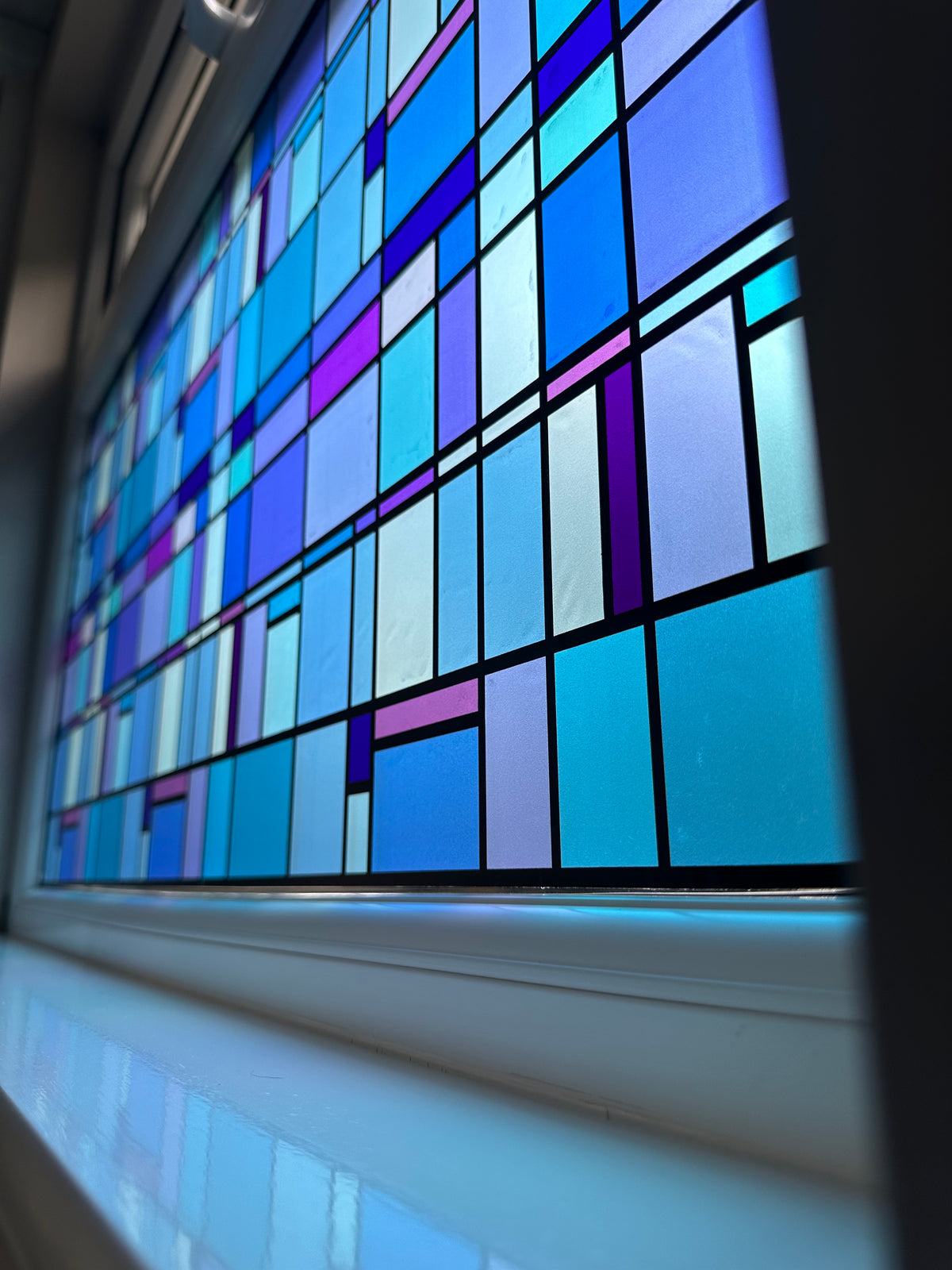 A close up of a window with stained glass effect window film applied. The film is a mosaic featuring shades of blue and purple. The film delivers a decorative upgrade and privacy.