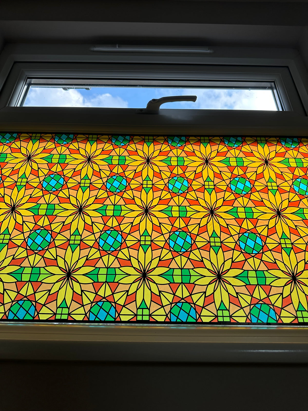 A colourful stained glass window panel with a repeating geometric star pattern in yellow, green, blue, and red, installed on a window in a residential property for decoration and privacy.