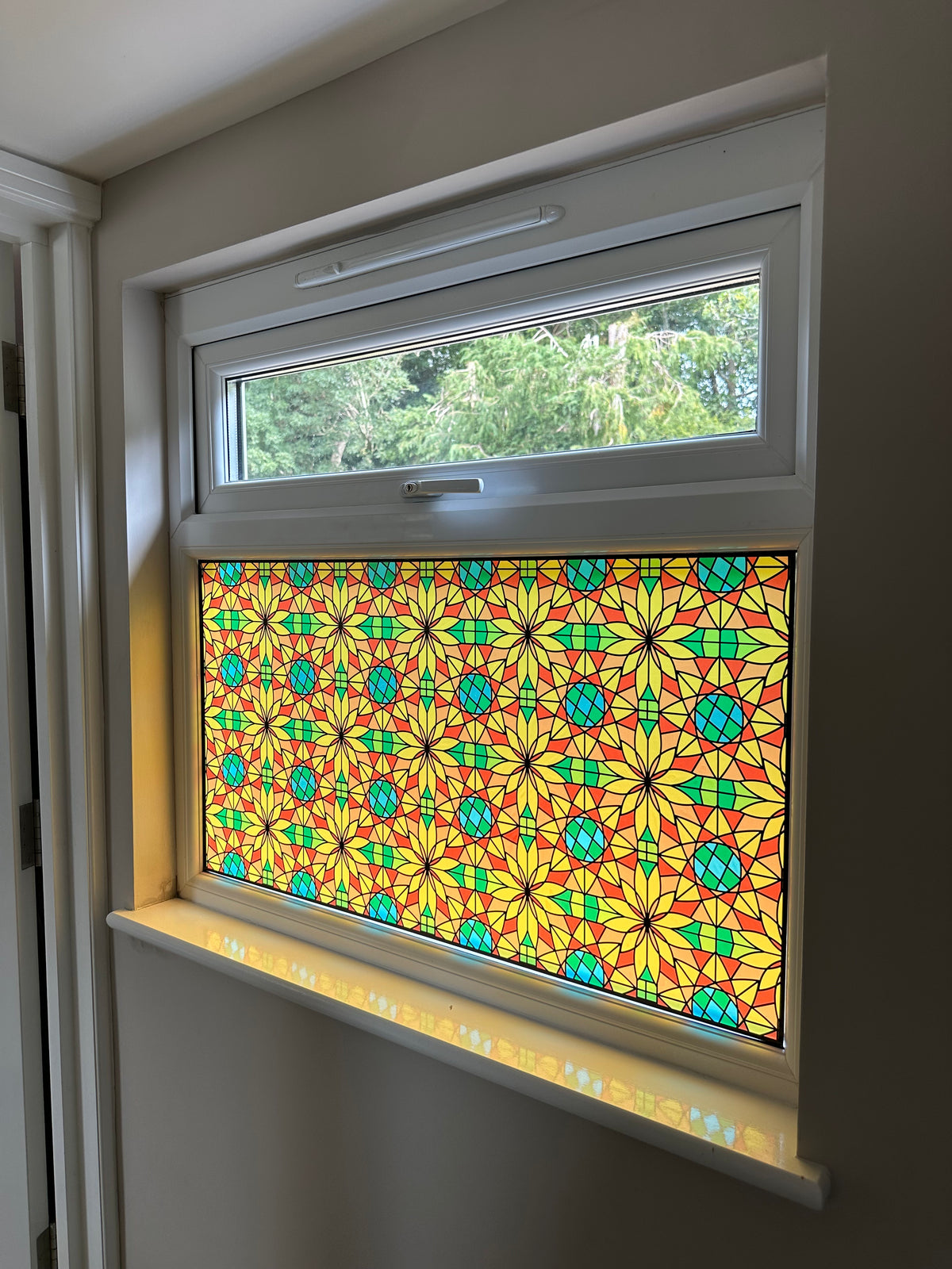 A decorative stained glass window panel with a colourful geometric star pattern, installed in a white uPVC window above a white ledge with colourful shadows cast onto the windowsill.