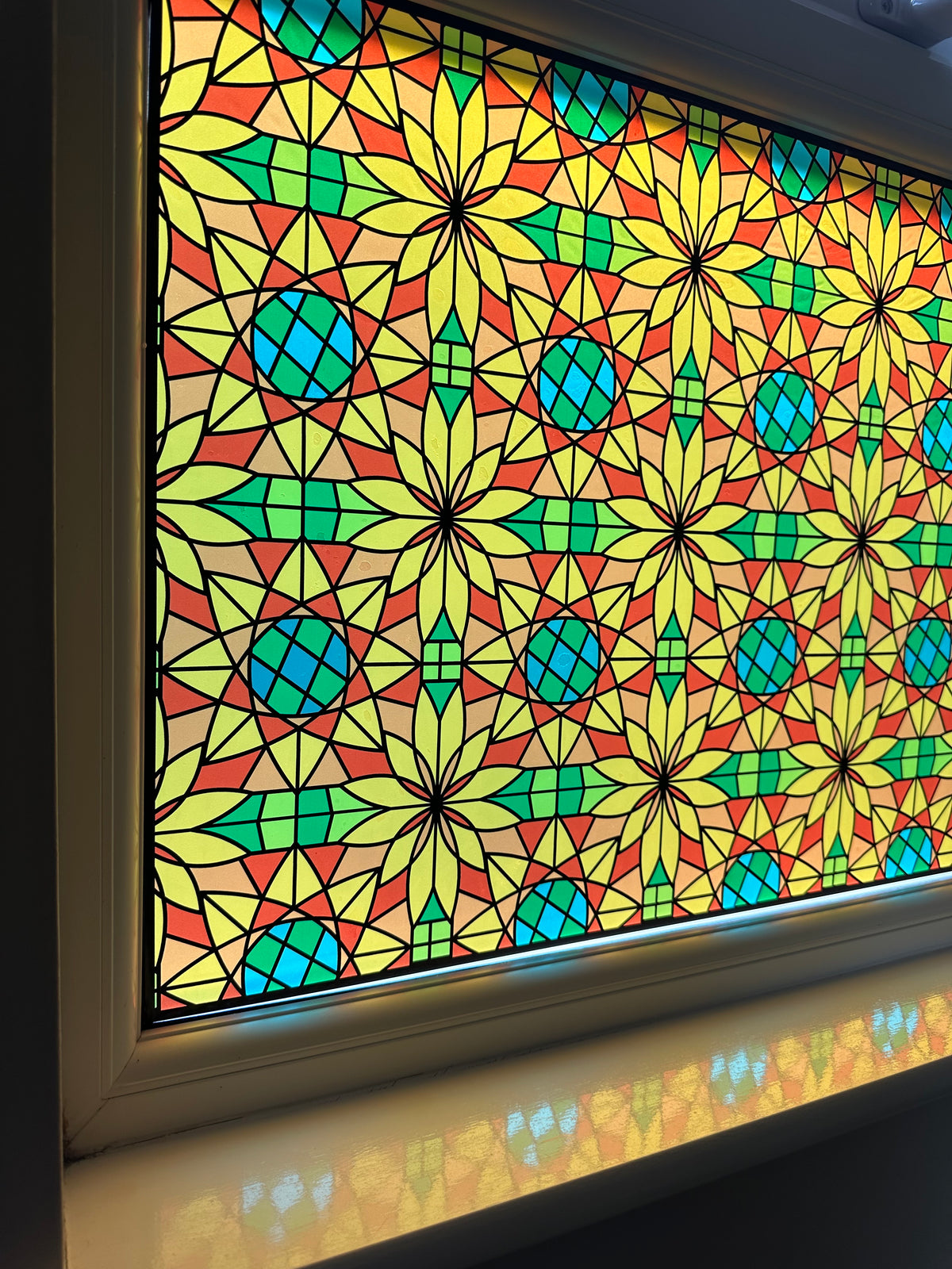 A close-up of a colorful stained-glass-style panel with repeating geometric star and circle motifs in yellow, green, teal, red, and beige, set in a window frame above a windowsill, with colourful shadows on the sill.