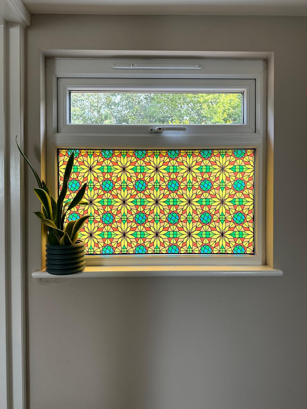 A decorative stained glass window panel with a colourful geometric pattern in yellow, green, and blue, installed on a white-framed window above a white windowsill with a small potted plant.