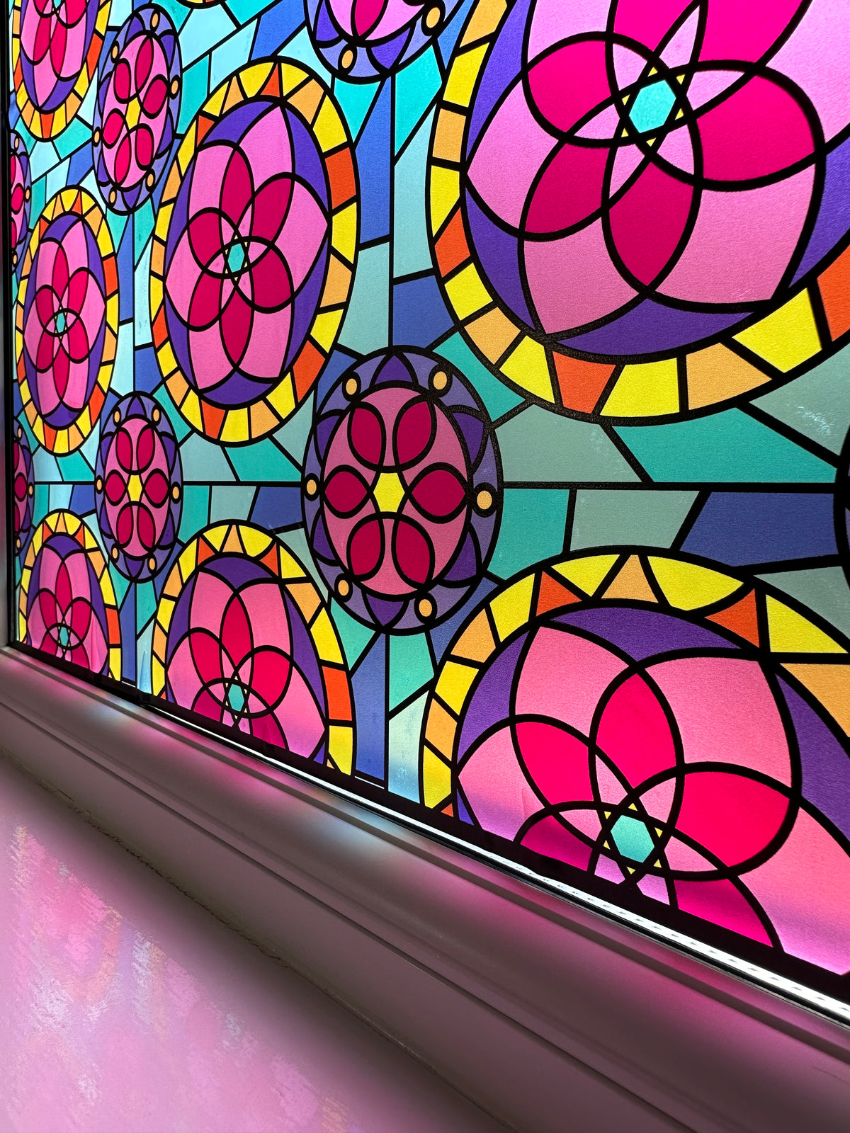 A close-up of a colorful stained-glass window design with repeating pink, magenta, purple, yellow, and teal floral and geometric motifs separated by dark lead lines.