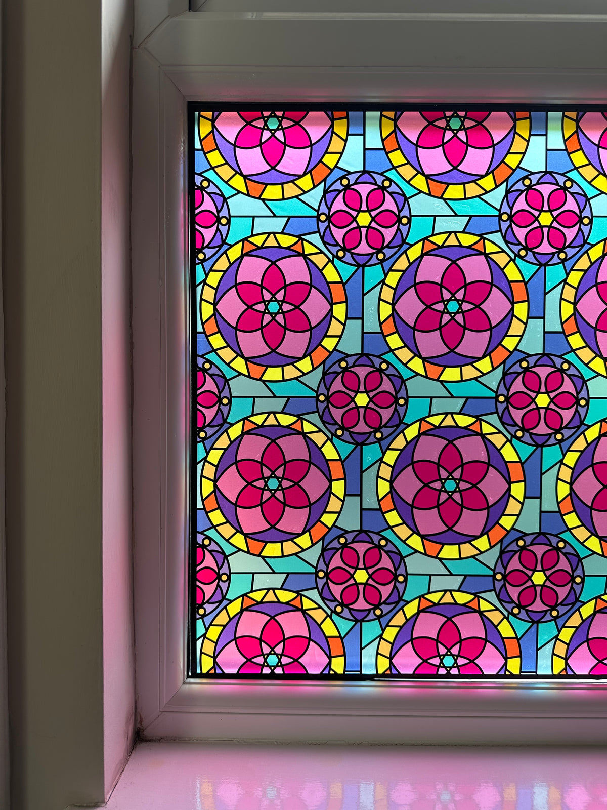 A close-up of a colorful stained-glass-style window panel with repeating circular floral motifs in pink, purple, yellow, and turquoise, applied to a window in a house for decor and privacy.