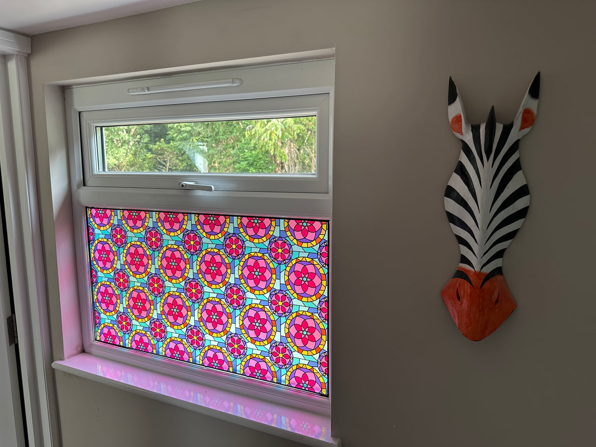 A colorful stained glass window panel with a repeating mandala pattern in pink, red, yellow, and blue, installed on a window in a residential property for decor and privacy.