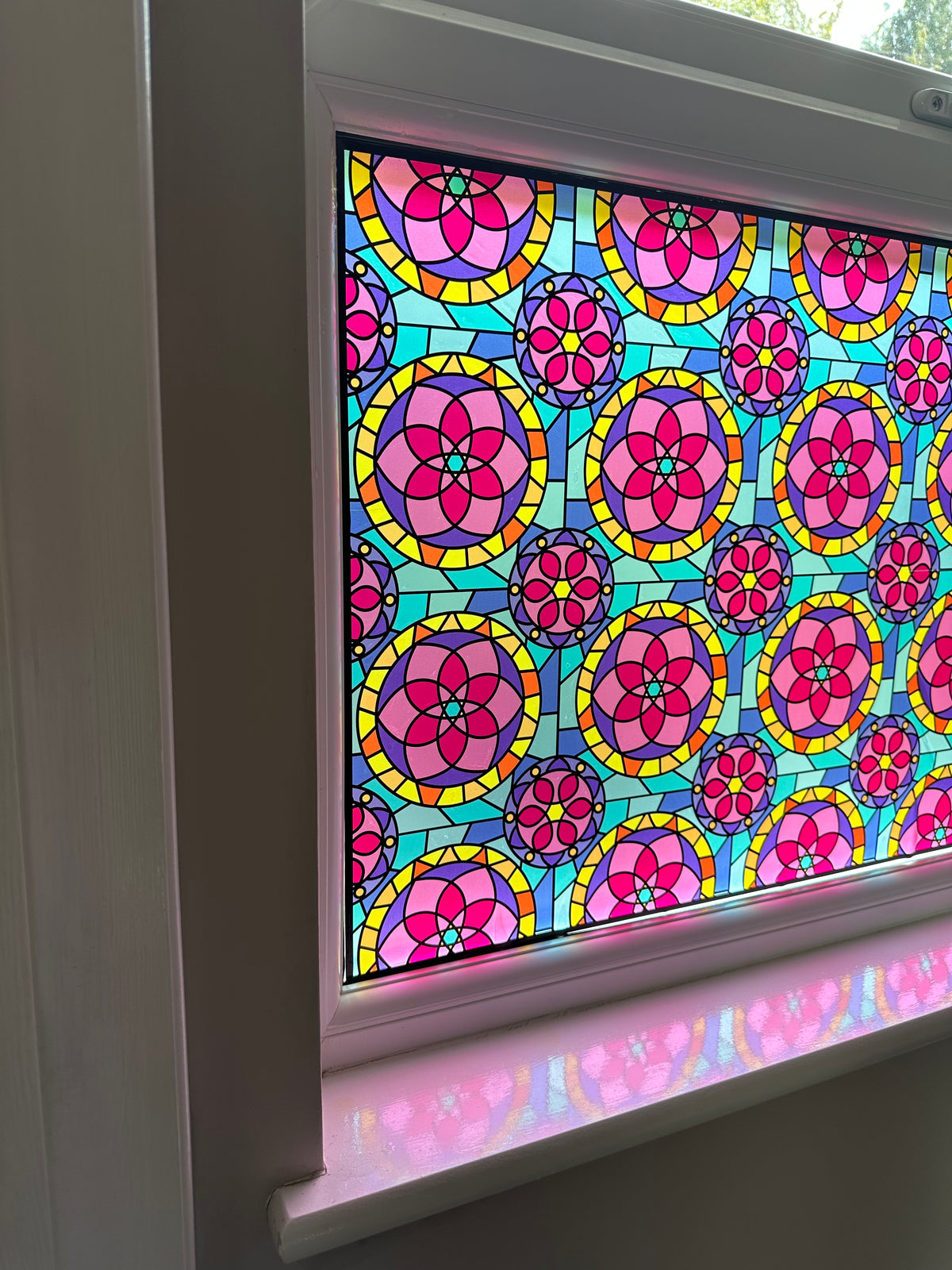 A close-up of a stained glass window panel featuring a repeating pattern of pink and yellow circular floral medallions on a turquoise background, set within a white window frame.