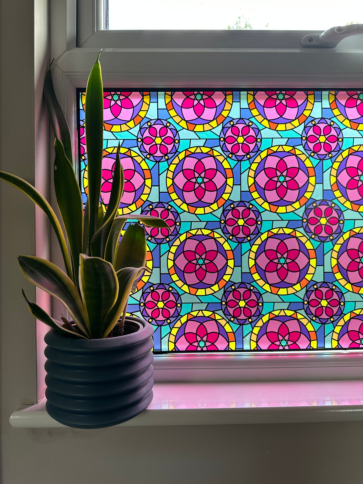 A colorful stained glass window with a repeating pink, yellow, and blue floral pattern, shown on a white window frame with a pottedplant on the sill.