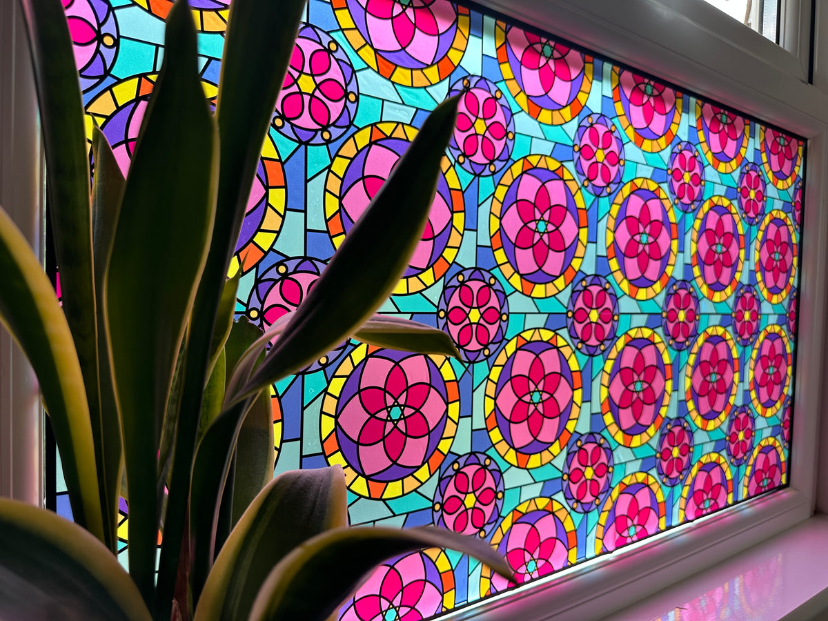 A large stained-glass style window panel with repeating pink, yellow, and blue floral circular motifs, shown installed on a window with a houseplant in the foreground.