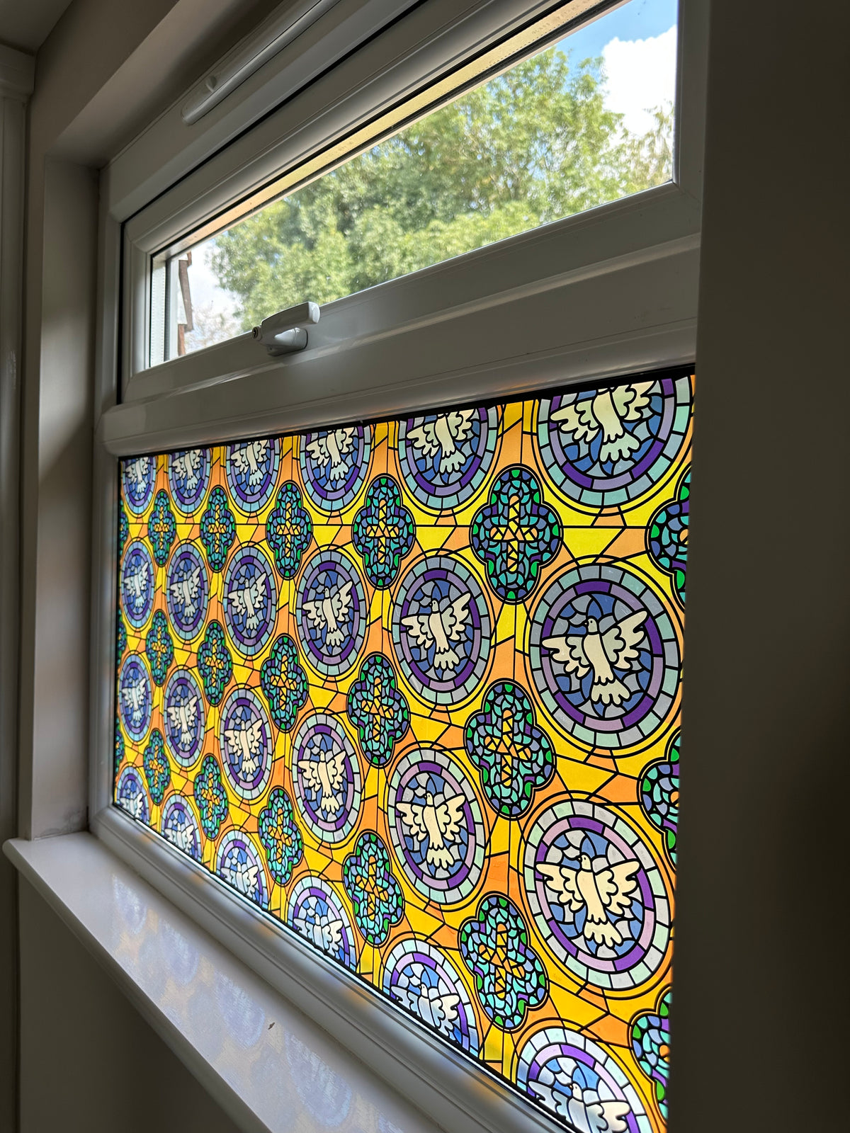 A close-up of a window with a decorative stained-glass panel featuring repeating circular medallions in yellow, blue, purple, green, and white, with outdoor greenery visible above the lower window pane.