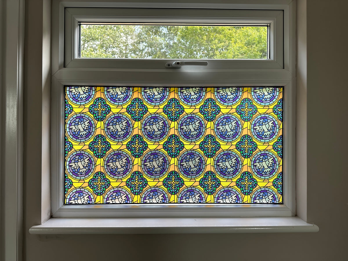 A decorative stained glass window panel with a repeating blue, yellow, and white mosaic style pattern, installed in a white-framed window.