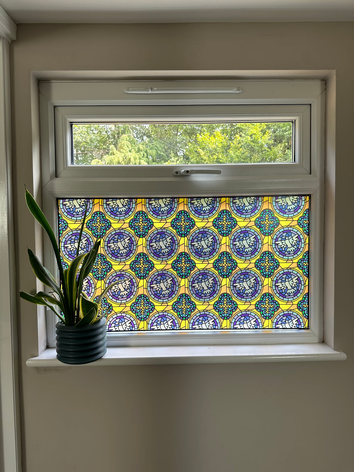 A decorative stained glass window panel with a repeating blue, yellow, and white geometric pattern, installed in a white casement window. A small potted green plant sits on the white window sill, and trees are visible outside.
