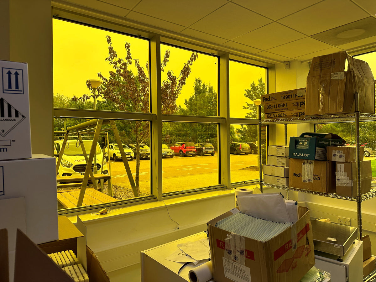 Storage area with boxes and shelves near a window showing an outdoor scene, Amber 81 UV protection window film has been applied to the glazing, giving a yellow hue to the scene. 