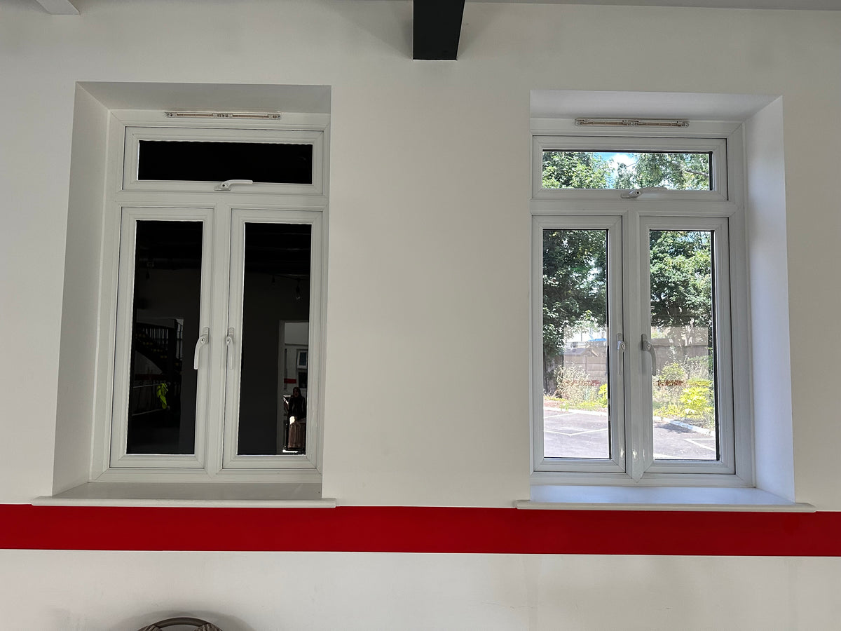 Two windows set into a white wall, the window on the left has blackout privacy window film applied to each of the three panes, while the window on the right allows a view out into a carpark.