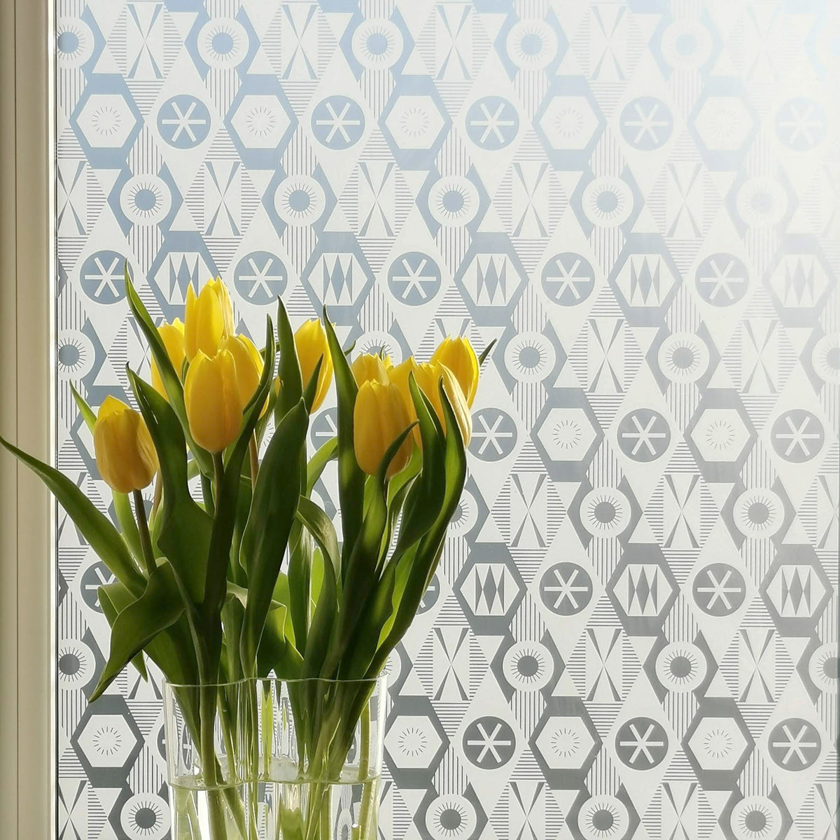 Close-up of a window with printed window film applied to it. The film features a repeated geometric design and there is a bunch of bright yellow daffodils on the window ledge. 