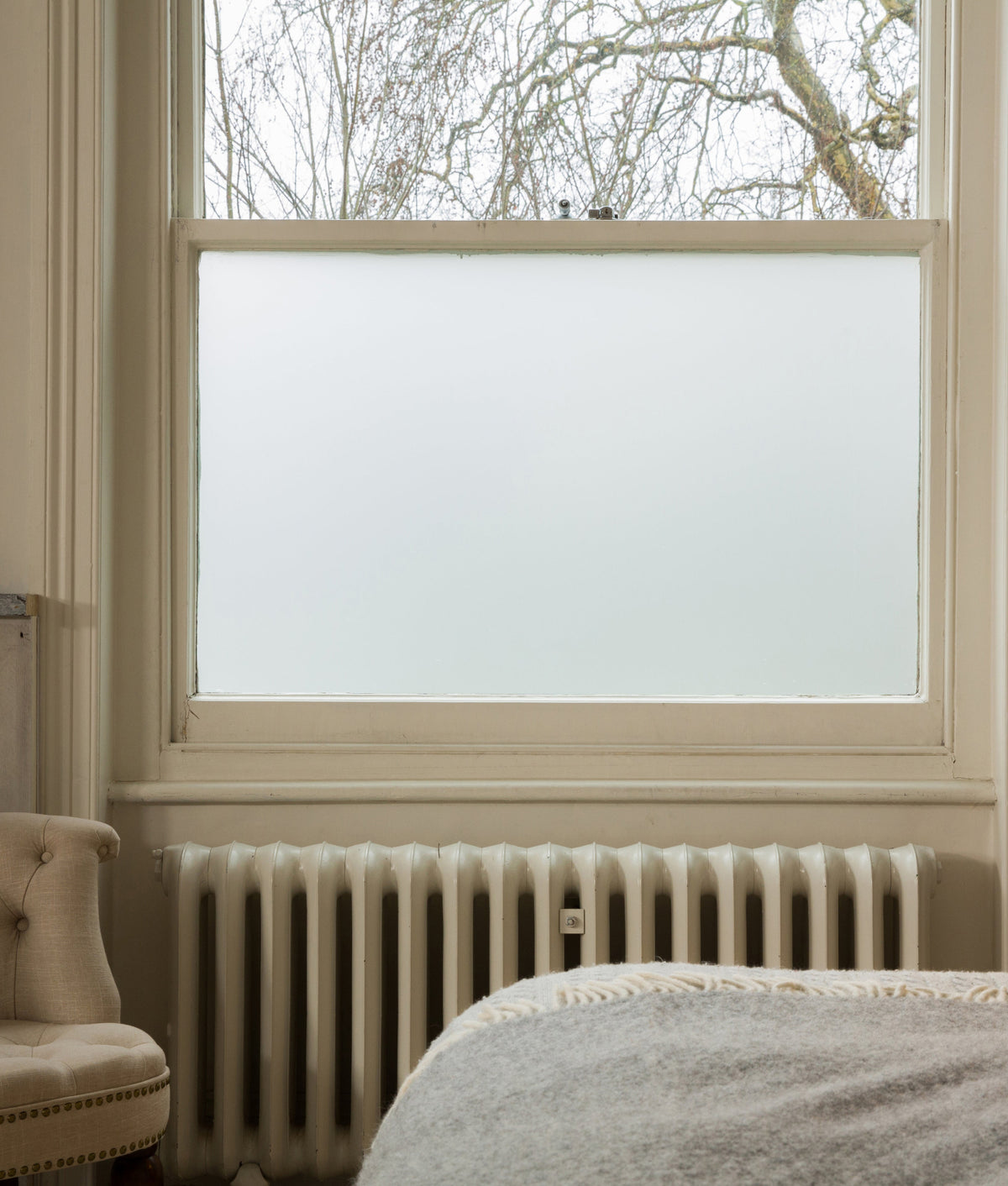 Frosted window film applied to a large window in a room with a radiator and chair for privacy.