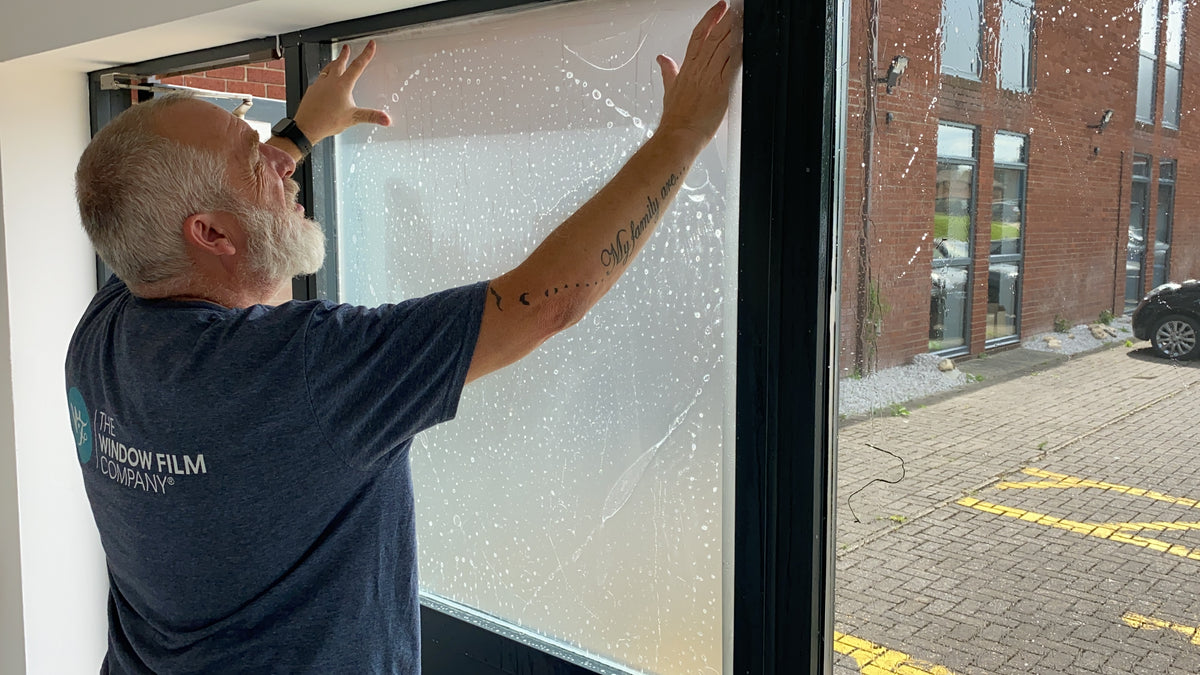 An installer from The Window Film Company fitting a frosted window film to the glass of an office building.