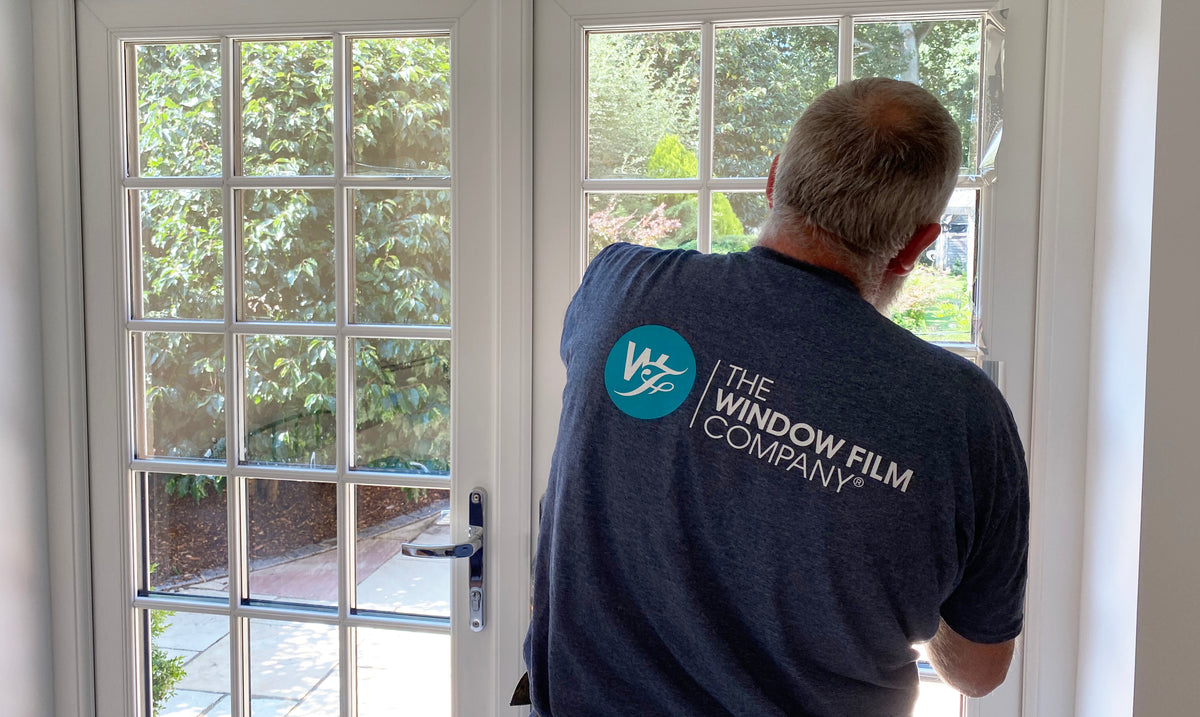 Person applying window film to a glass door with 'The Window Film Company' branding on the back of the t-shirt.