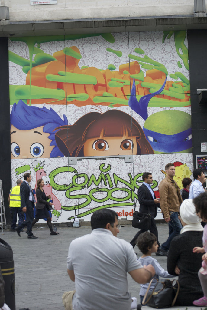 Printed mural of cartoon characters with 'Coming Soon' text on a building wall, people walking by.