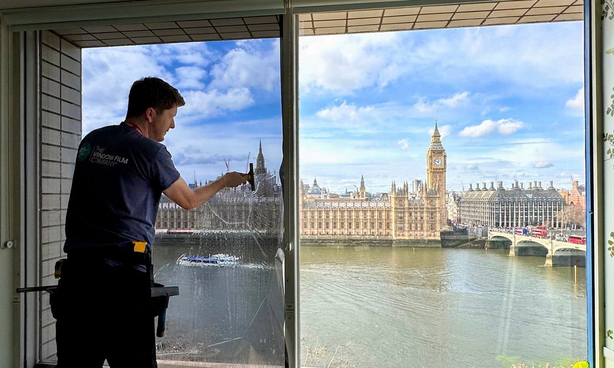 A window film installer fitting Optiview 15 solar control window film to a window in London overlooking Big Ben.  One window has film and the other doesn't for a comparison.