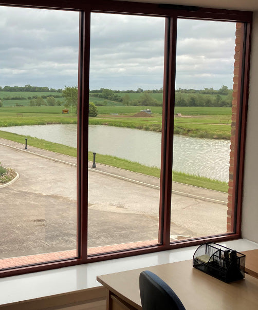 An internal view of a large window, looking out over green fields and a lake. The glass has solar control window film applied, giving the appearance of the glass a slight tinted finish.
