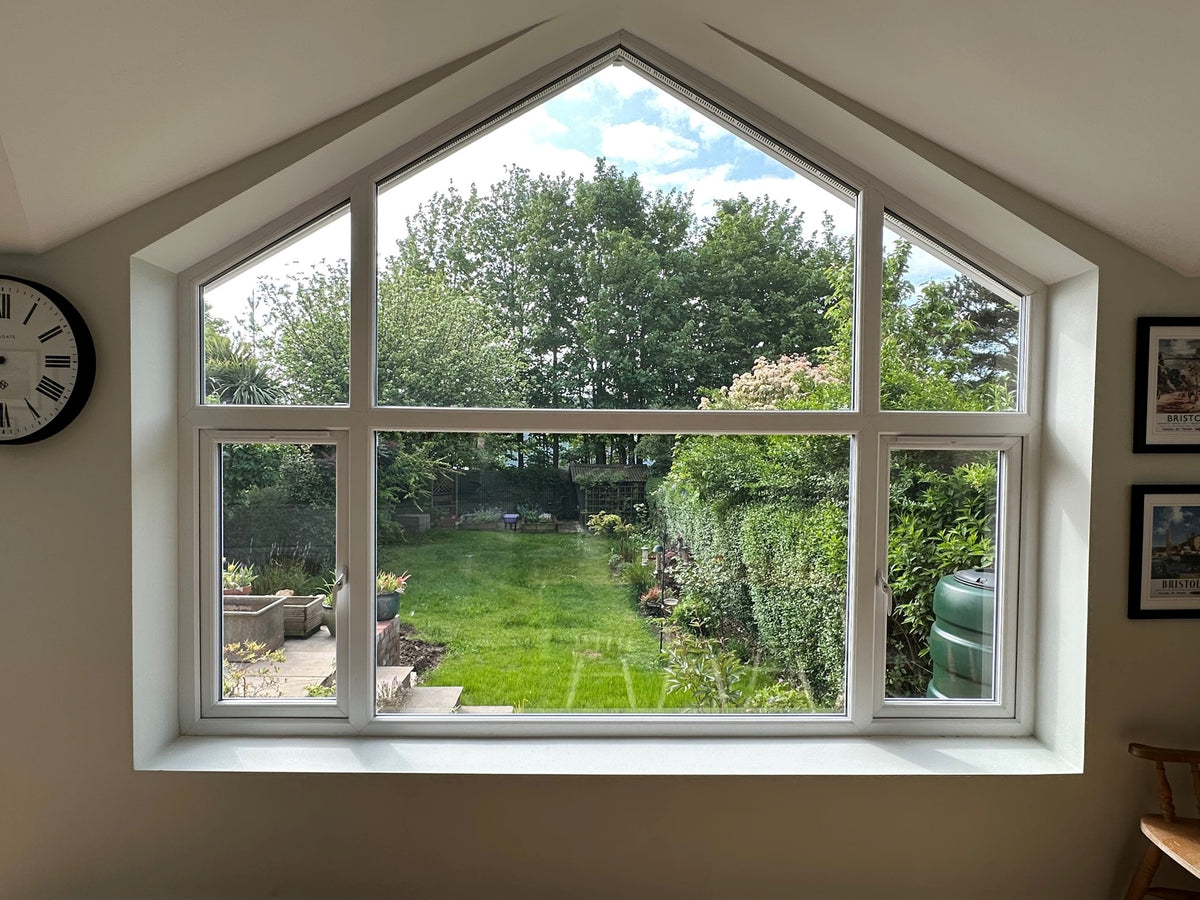 Large window with heat and glare reduction window film applied to it, with a view of a garden and trees, with a clock and framed pictures on the wall.