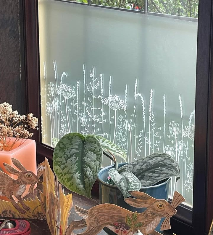 A window with frosted window film with a white printed nature-inspired pattern applied, with potted plants on the window ledge in the foreground. 