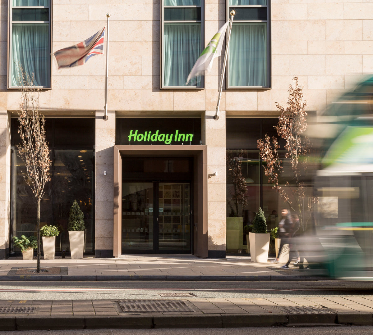 The entrance to a large Holiday inn hotel in Manchester. A bus is speeding past and safety window film has been applied to the glazing.  