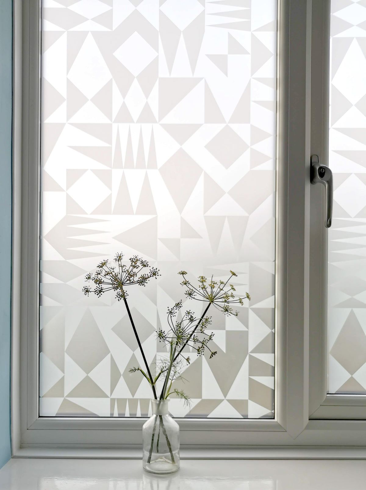 Window with geometric patterned film and a vase with flowers on a white surface.