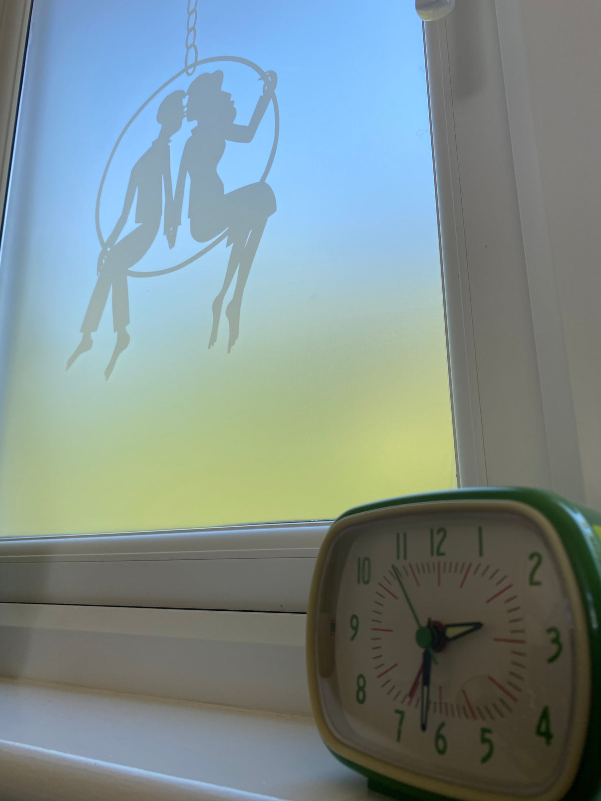 Window with a white frame and window ledge, and Rob Ryan frosted window film applied to the glass. There is a green clock on the ledge in the foreground. 