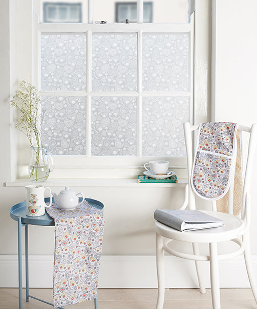 Room with white walls, a chair and bistro table in the foreground and a window with white window frames. The glass has had Birdie Folk window film by helen Russell applied to it.
